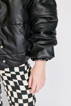 Cargar imagen en el visor de la galería, Chaqueta Puffer de mujer. Diseño alcochado en cuero sintético. Tejido técnico repelente al agua. Cuero Vegano. Cuello subido y manga larga con puños elásticos. Bolsillos laterales. Cierre frontal con botones a presión ocultos por la solapa. Forro interior con estampado de Flint. Disponible en color Café y Negro. Producto 100% hecho en Colombia.