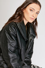 Cargar imagen en el visor de la galería, Chaqueta Puffer de mujer. Diseño alcochado en cuero sintético. Tejido técnico repelente al agua. Cuero Vegano. Cuello subido y manga larga con puños elásticos. Bolsillos laterales. Cierre frontal con botones a presión ocultos por la solapa. Forro interior con estampado de Flint. Disponible en color Café y Negro. Producto 100% hecho en Colombia.